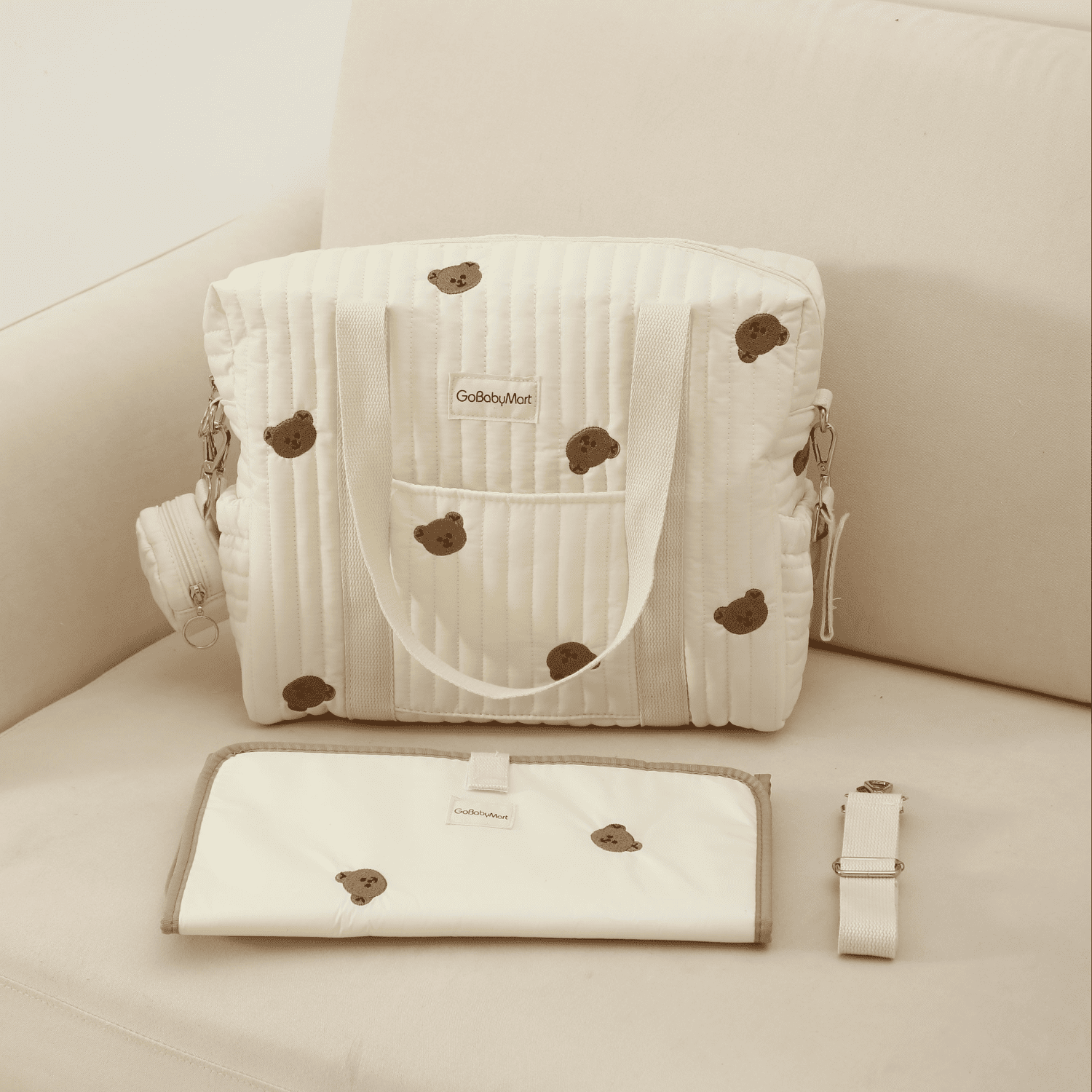 An off-white quilted diaper bag with brown teddy bear prints, shown with its matching flat storage pouch and strap on a beige sofa. The brand name 'GodbyMiki' is visible on the labels. #Color_Beige Bears